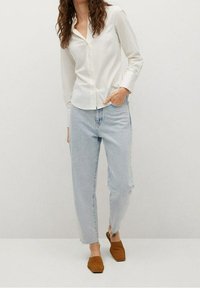 Chemise blanche à boutons, jean taille haute bleu clair, et chaussures marron sans lacets. Tissu lisse, coupe décontractée, et détails d'ourlet effiloché.