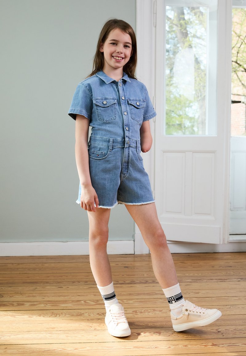 Salopette en denim à manches courtes et poches avant, couleur bleu clair, ourlet effiloché ; associée à des baskets blanches et des chaussettes rayées.