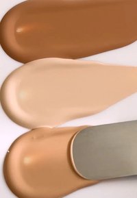 Tre kremfoundation-skvott på en hvit overflate: mørk brun, lys beige og ferskenbeige, med en metallspatel for påføring.