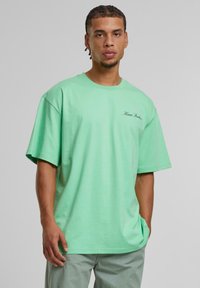 Mint green oversized t-shirt with short sleeves, round neckline, and black logo stitching on the left chest. Soft cotton material.