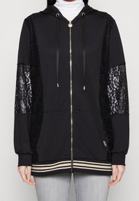 Černá mikina na zip s krajkovými rukávy, kapucí a zlatými kovovými doplňky. Má pruhovaný lem a texturovanou látku pro kontrast.