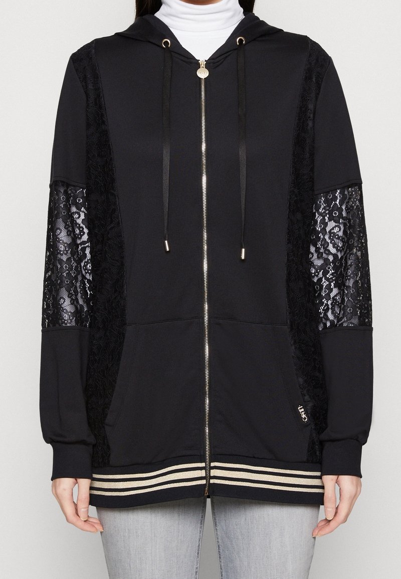 Černá mikina na zip s krajkovými rukávy, kapucí a zlatými kovovými doplňky. Má pruhovaný lem a texturovanou látku pro kontrast.