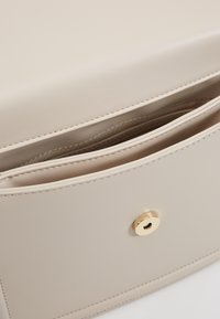 Beige konstläderväska med en enkel design, framträdande guldfärgad hårdvara och en öppen ficka med en slät yta och rena sömmar.