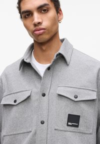 Camisa chaqueta de mezcla de lana gris con dos bolsillos en el pecho que presentan botones a presión negros y un parche con logotipo negro en el bolsillo izquierdo.