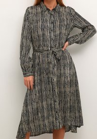 Frau in einem schwarz-beigen gestreiften Hemdblusenkleid mit langen Ärmeln und einem gebundenen Taillengürtel, die mit einer Hand auf der Hüfte steht.