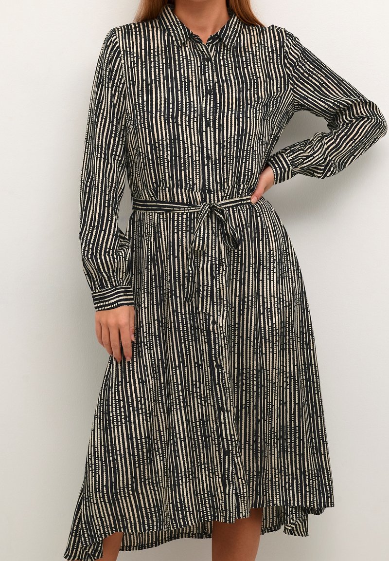 Frau in einem schwarz-beigen gestreiften Hemdblusenkleid mit langen Ärmeln und einem gebundenen Taillengürtel, die mit einer Hand auf der Hüfte steht.