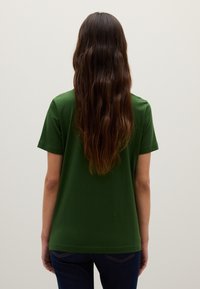 Grünes Baumwoll-T-Shirt mit kurzen Ärmeln, lockerer Passform und abgerundetem Saum, von hinten betrachtet. Dunkelblaue Jeans vervollständigen das Outfit.