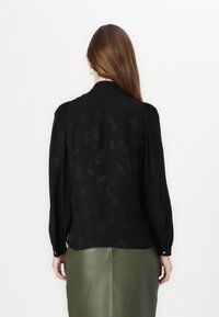 Svart bluse med lange ermer, subtil blomster- mønster, myk stoff, og kragehals. Puffede ermer og knappe mansjetter gir detaljer.