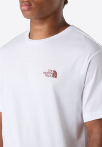 Muž v bíl�ém tričku s kulatým výstřihem, na kterém je hnědé logo The North Face na levé části hrudi, záběr zkrácený na krk a ramena.