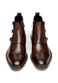 Botas de cuero marrón hasta el tobillo con un acabado pulido, que presentan tres adornos de hebilla, punta afilada y una lengüeta en la parte trasera para facilitar el uso.