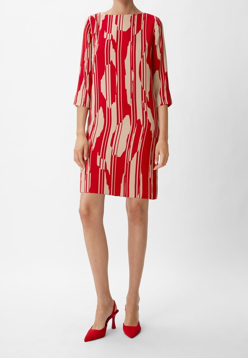 Femme portant une robe rouge et beige à motifs, longueur genoux, manches trois-quarts, et des escarpins rouges à bout pointu sur fond blanc.