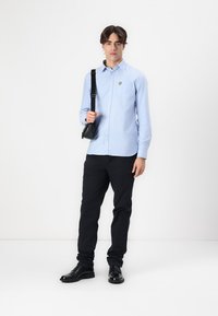 Chemise bleu clair à boutons avec un petit logo, portée avec un pantalon noir et des chaussures noires cirées. Le look est complété par un sac bandoulière noir.