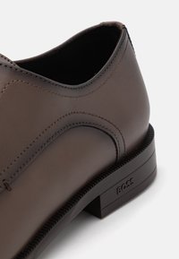Scarpa in pelle marrone con superficie liscia, dettagli cuciti e tacco basso. Presenta un logo sul tacco e una sottile linea curva nel design.