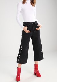 Svarta cropped-byxor med grommetdetaljer på sidorna, i kombination med en vit långärmad topp och glänsande röda ankelstövlar.