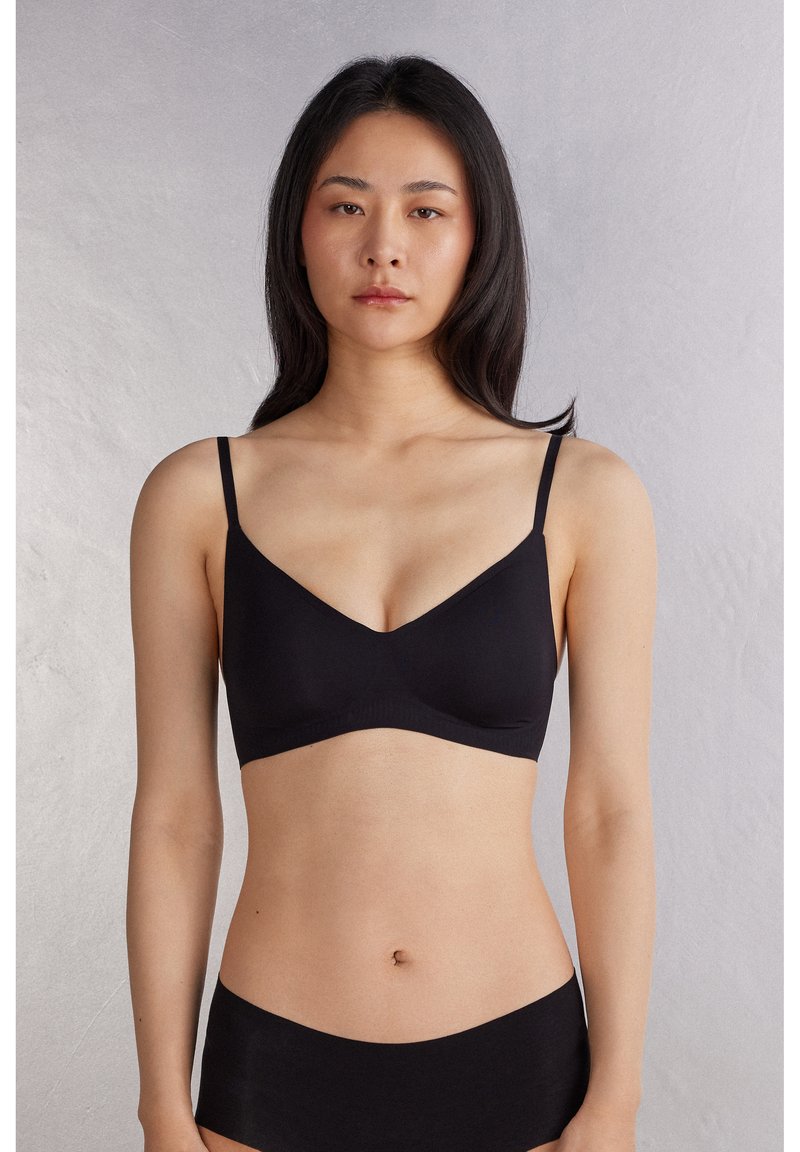 Joven mujer con cabello largo y negro, vestida con un sujetador suave negro y ropa interior negra a juego, de pie frente a un fondo liso gris claro.