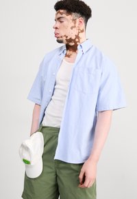 Vaaleansininen lyhythihainen paita, napitettavalla tyylillä, jossa on yksi tasku. Yhdistetty valkoisen tanktopin ja vihreiden shortsien kanssa, pitäen valkoista lippistä.
