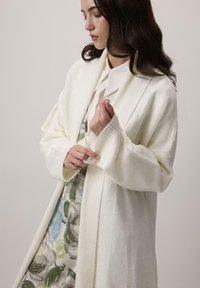 Biely pletený kardigán s otvoreným predkom, rebrovanými manžetami a mäkkou textúrou, prehodený cez vzorované kvietkové šaty s zelenými a modrými akcentmi.