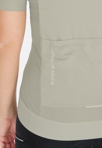 Lähikuva henkilöstä, joka pukeutunut vaaleanharmaaseen, teksturoituun pyöräilypaitaan, jossa on takataskut ja "ENDURANCE"-teksti, yhdistettynä mustiin shortseihin.