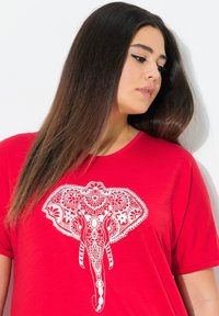 T-shirt rouge arborant un grand graphisme d'éléphant blanc avec des motifs complexes. Manches courtes et coupe décontractée.