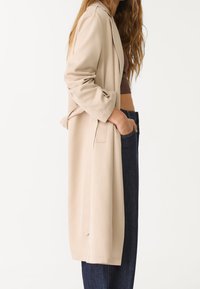 Beżowy długi płaszcz o luźnym kroju, wykonany z lekkiego materiału, z kołnierzykiem z wycięciem, bocznymi kieszeniami i podwiniętymi rękawami.