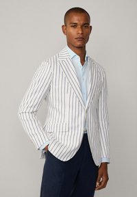 Blazer rayé blanc et marine, en tissu léger. Les caractéristiques notables comprennent deux poches avant et des accents de boutons dorés.