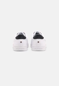 White sneakers with a black heel accent, flat sole, smooth leather texture, and "HILFIGER" branding on the back. Minimalistic design.