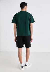 Grønn bomull t-skjorte, kortermet, avslappet passform, kombinert med sorte shorts. Modellen har på seg hvite sneakers med blå detaljer og hvite sokker.