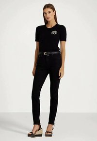 Mulher vestindo uma blusa preta de mangas curtas com logotipo branco, calças justas pretas, cinto preto e sandálias de plataforma pretas, de pé contra um fundo neutro.