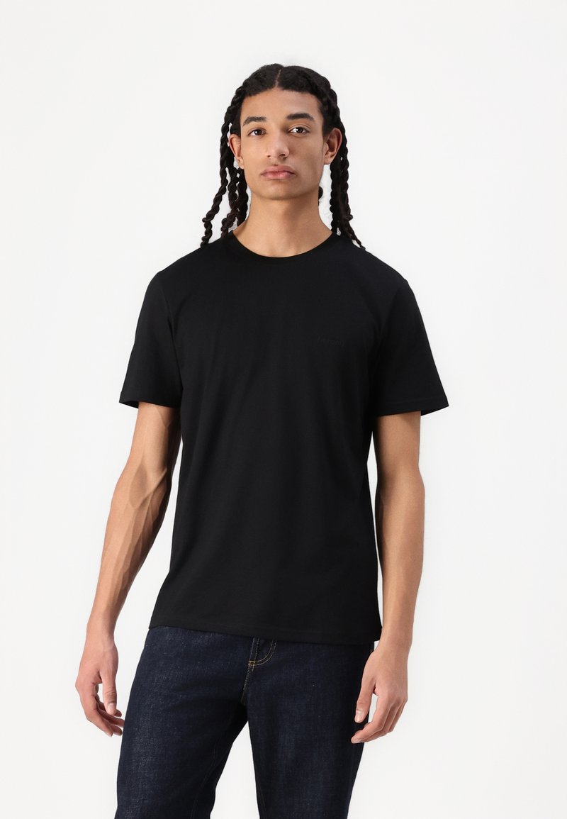 Sort bomulds t-shirt med korte ærmer, rund hals og et diskret logo på brystet. Modellen har blå jeans på.