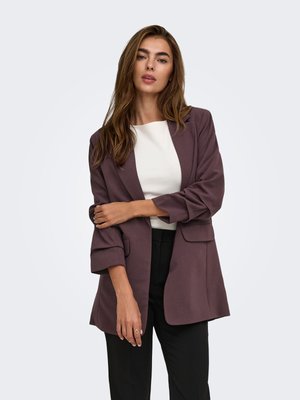 Mujer con cabello largo y castaño que lleva una blazer morada sobre una camiseta blanca y pantalones negros, de pie con un brazo cruzado sobre su cuerpo.