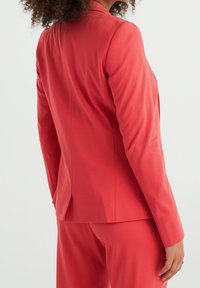 Korallfarbener Blazer mit tailliertem Schnitt, glatter Textur und einem Schlitz hinten. Mit passender Hose, sichtbaren Nähten und langen Ärmeln.