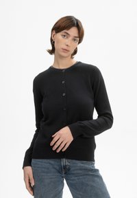 Zwarte gebreide cardigan met een ronde hals, knoopsluiting en geribde manchetten. Gecombineerd met lichtblauwe denim jeans.