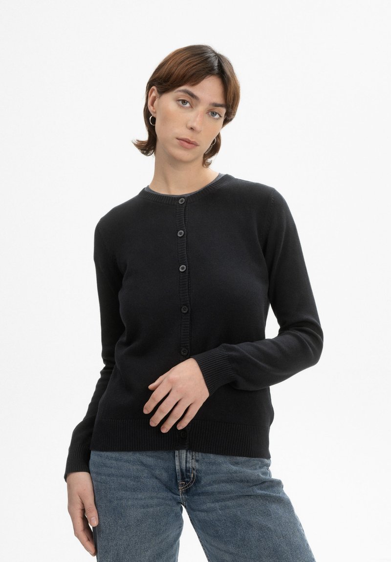 Zwarte gebreide cardigan met een ronde hals, knoopsluiting en geribde manchetten. Gecombineerd met lichtblauwe denim jeans.