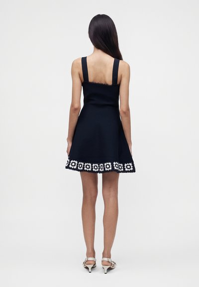 Femme aux longs cheveux foncés portant une robe noire sans manches avec un ourlet bordé de fleurs blanches et des sandales blanches à talons, debout, dos tourné.
