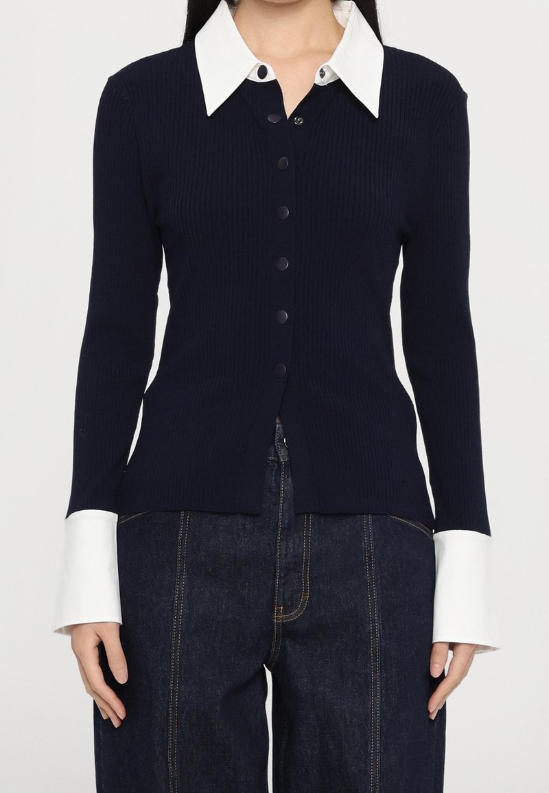Femme portant un cardigan côtelé bleu marine boutonné avec un col et des poignets blancs surdimensionnés, associé à un jean denim foncé avec des coutures visibles.