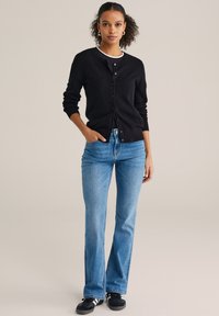 Fekete texturált kardigán elöl gombokkal, világoskék, bővülő szárú farmerrel párosítva. A modell fekete, fehér részletekkel díszített sportcipőt visel.