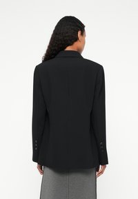 Blazer nero su misura con colletto a punta, maniche lunghe e quattro bottoni su ciascun polsino. Tessuto liscio con una silhouette strutturata.