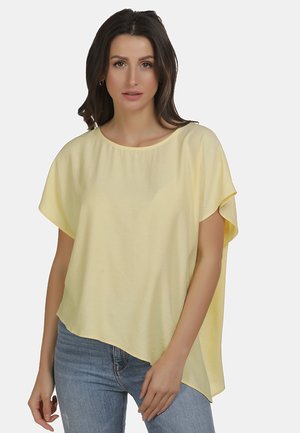 Jonge vrouw met donker haar die een losse lichtgele korte mouw blouse en blauwe spijkerbroek draagt, staand tegen een lichte achtergrond.