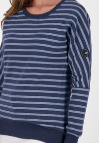 Woman wearing a navy and light blue horizontal striped long-sleeve shirt with a round neckline and a small circular logo on the sleeve.