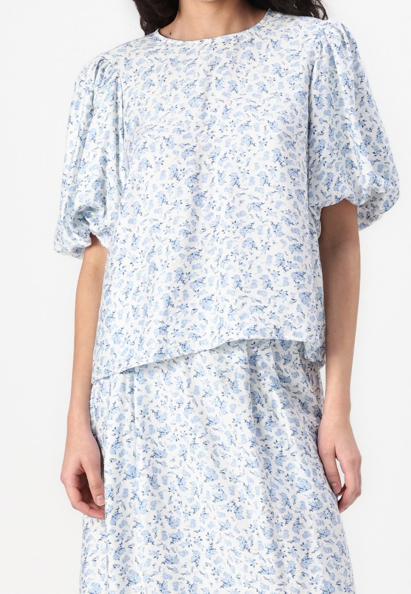 Femme portant un chemisier blanc et une jupe avec un délicat motif floral bleu et des manches courtes bouffantes.