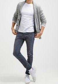Gray knitted cardigan with buttons, white t-shirt, slim-fit gray jeans, and white sneakers with black accents. White background.