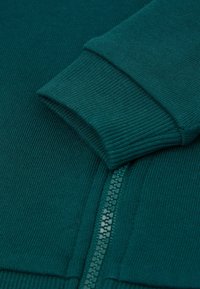 Pull en teal avec des poignets côtelés et une fermeture éclair, fabriqué à partir d'un tissu texturé. La fermeture éclair est un accent visible le long de la couture.