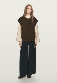 Brown oversized vest with button closure, worn over a light-colored long-sleeve top and paired with wide-leg black trousers. Smooth textures and simple design.