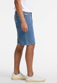 Shorts de denim en azul claro, con un corte recto, dobladillo deshilachado y bolsillos traseros. Combinados con zapatillas blancas, con tejido texturizado visible.