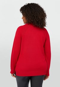 Czerwony sweter z długim rękawem wykonany z gładkiej dzianiny, z wysokim dekoltem i bocznymi rozcięciami na dolnym brzegu. Wygięty krój z tyłu.