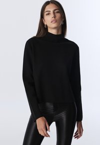 Mladá žena se rovnými hnědými vlasy, která má na sobě stříbrné kruhové náušnice, černý top s dlouhými rukávy a černé kožené kalhoty na bílém pozadí.