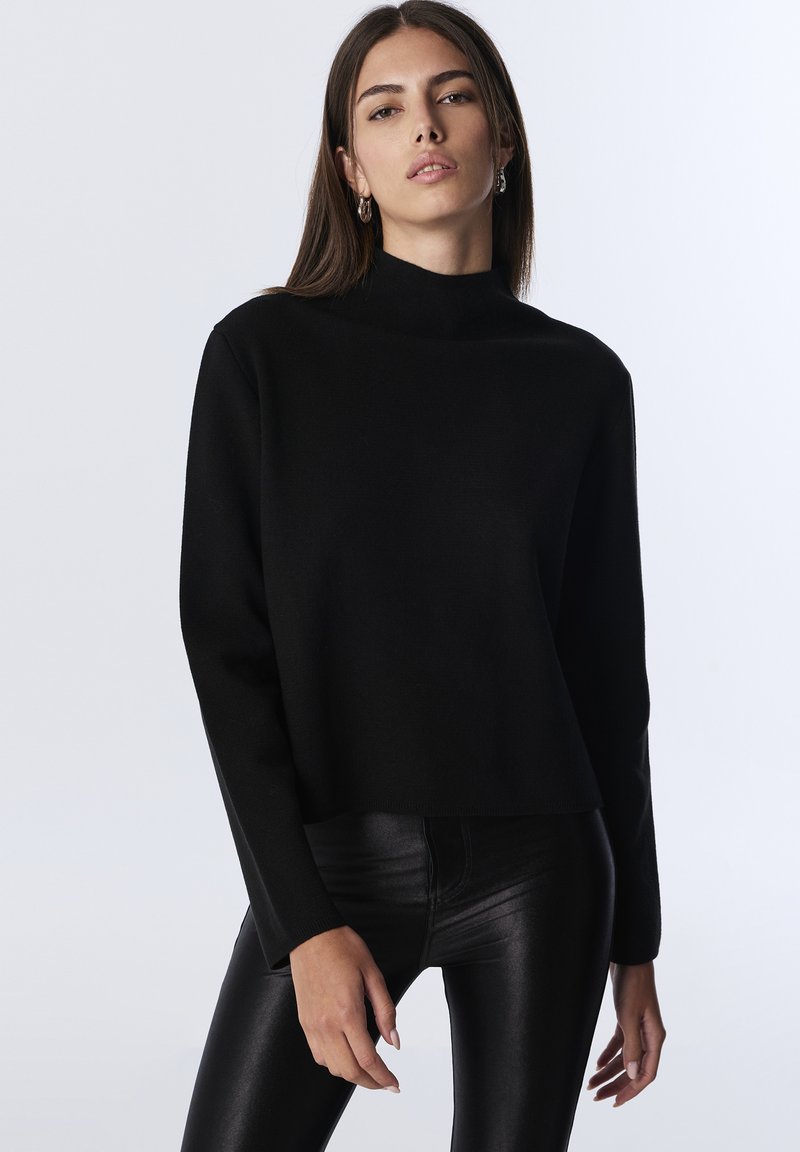 Mladá žena se rovnými hnědými vlasy, která má na sobě stříbrné kruhové náušnice, černý top s dlouhými rukávy a černé kožené kalhoty na bílém pozadí.