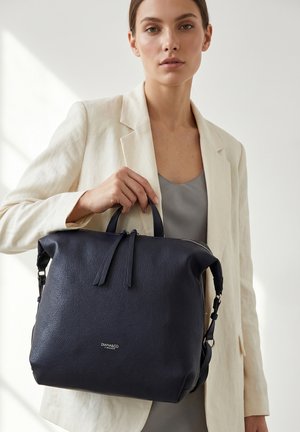 Femme en blazer beige tenant un grand sac à main en cuir noir avec des poignées supérieures et une fermeture éclair visible, de la marque "DIANA&CO FIRENZE."