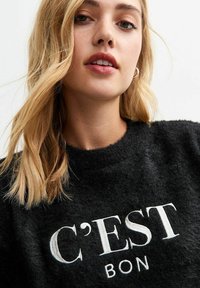 Maglione nero peloso con scritte bianche "C’EST BON", scollatura rotonda e tessuto testurizzato. Il modello ha capelli biondi ondulati e trucco sobrio.