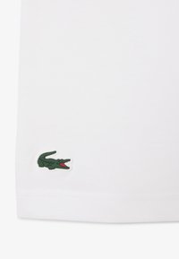 Logo de cocodrilo verde con boca roja bordado en la esquina de una prenda de tela blanca.